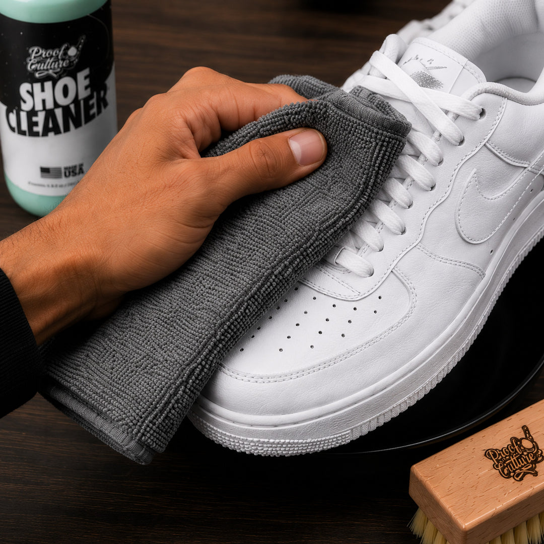 Hand buffing a white Air Force 1 sneaker with the grey microfiber towel, Proof Culture Shoe Cleaner and brush nearby