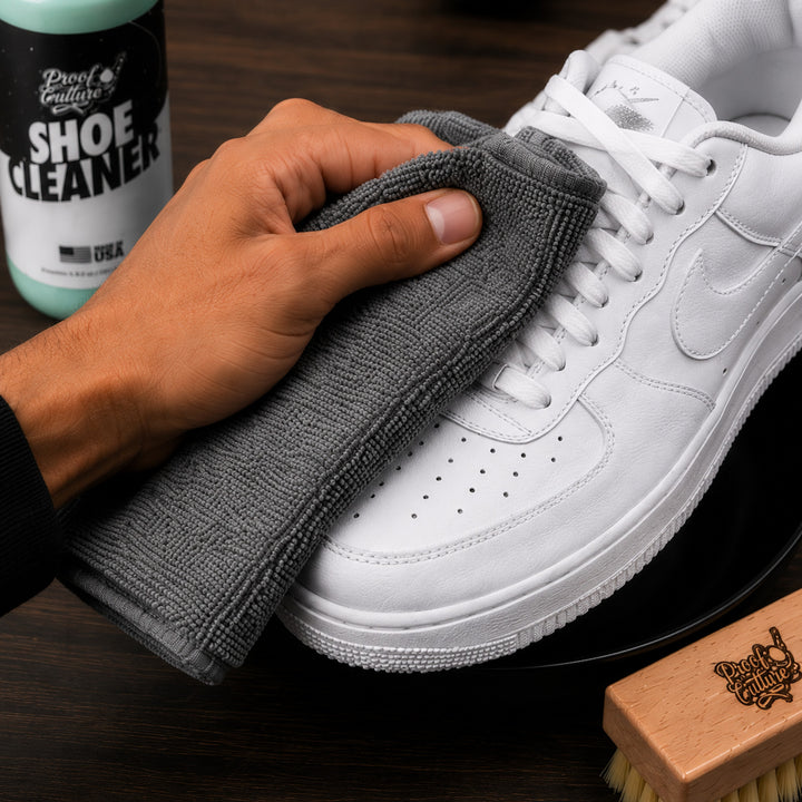 Hand buffing a white Air Force 1 sneaker with the grey microfiber towel, Proof Culture Shoe Cleaner and brush nearby