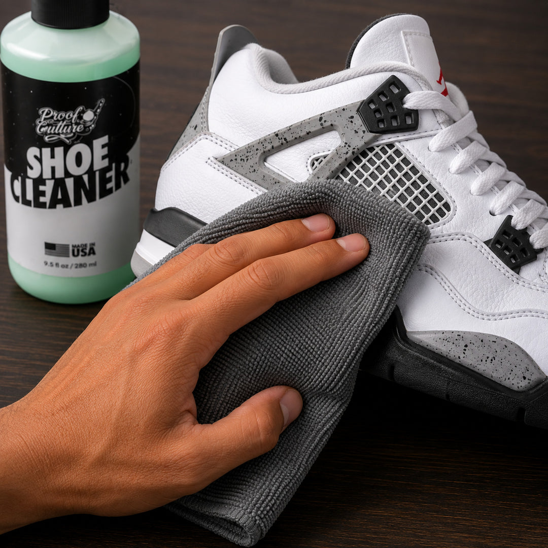 Hand wiping a white and grey Jordan 4 sneaker with the grey microfiber towel, Proof Culture Shoe Cleaner bottle in the background