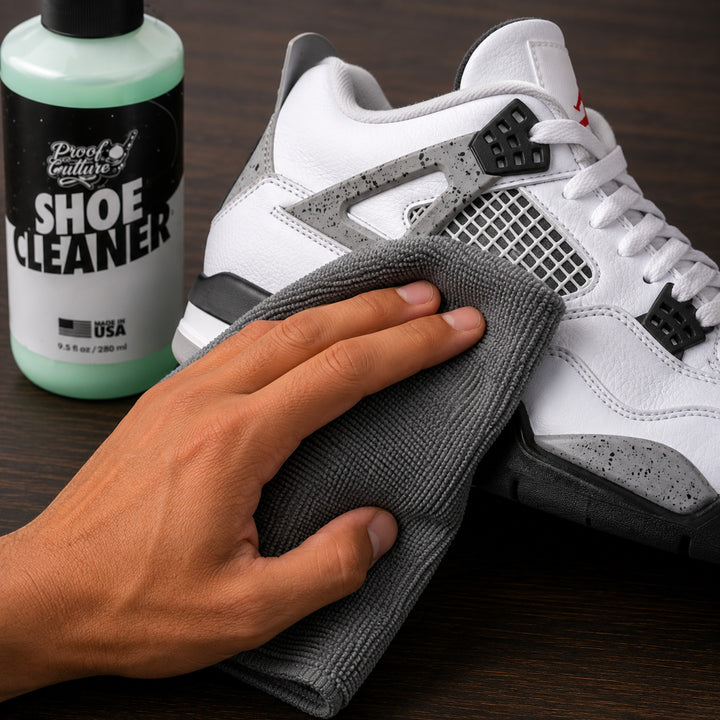 Hand wiping a white and grey Jordan 4 sneaker with the grey microfiber towel, Proof Culture Shoe Cleaner bottle in the background